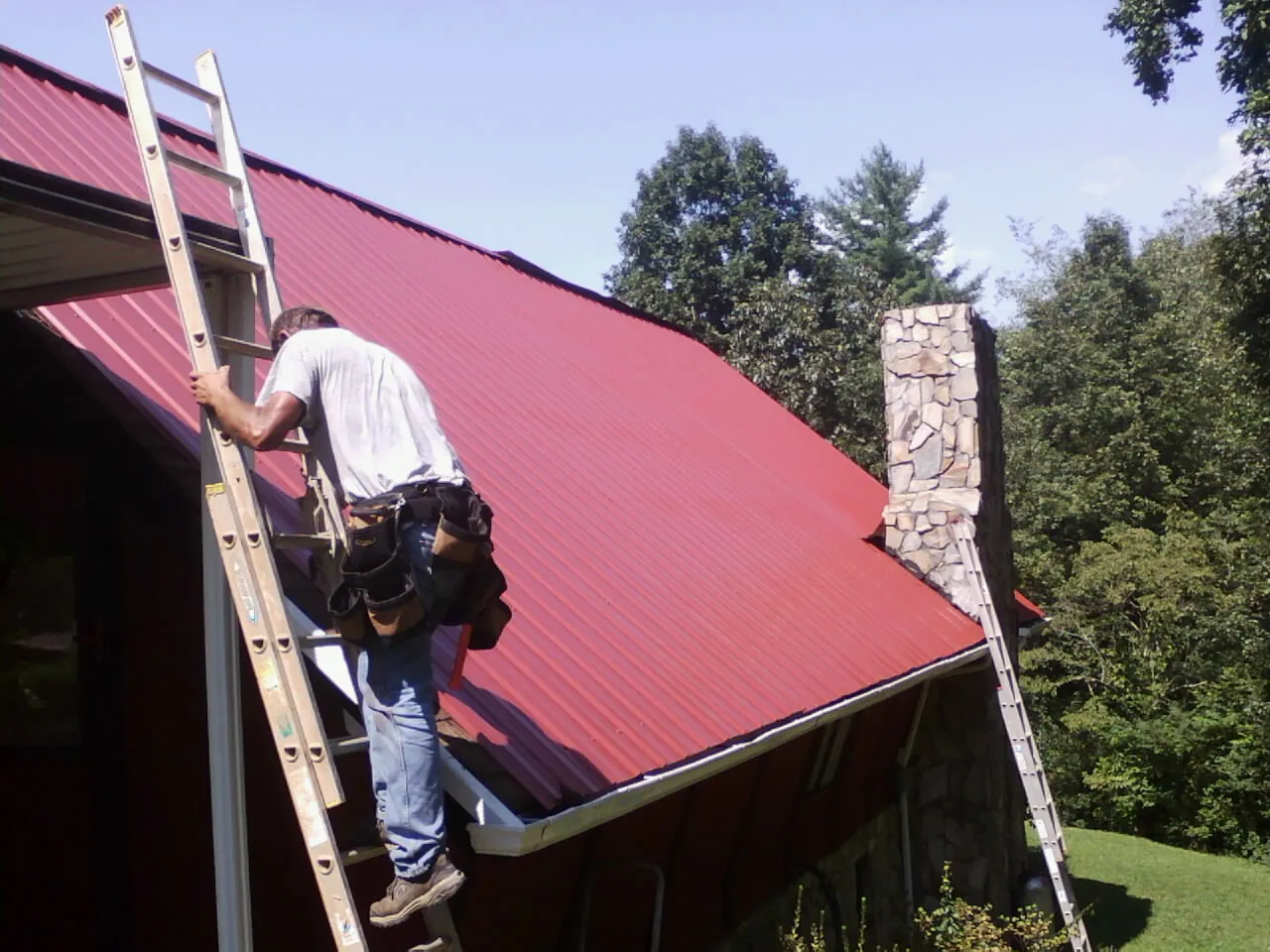 Roofer on a ladder beside a red metal roof and stone chimney — Soffit & Fascia Repair by Roofing Brothers in Milton Freewater